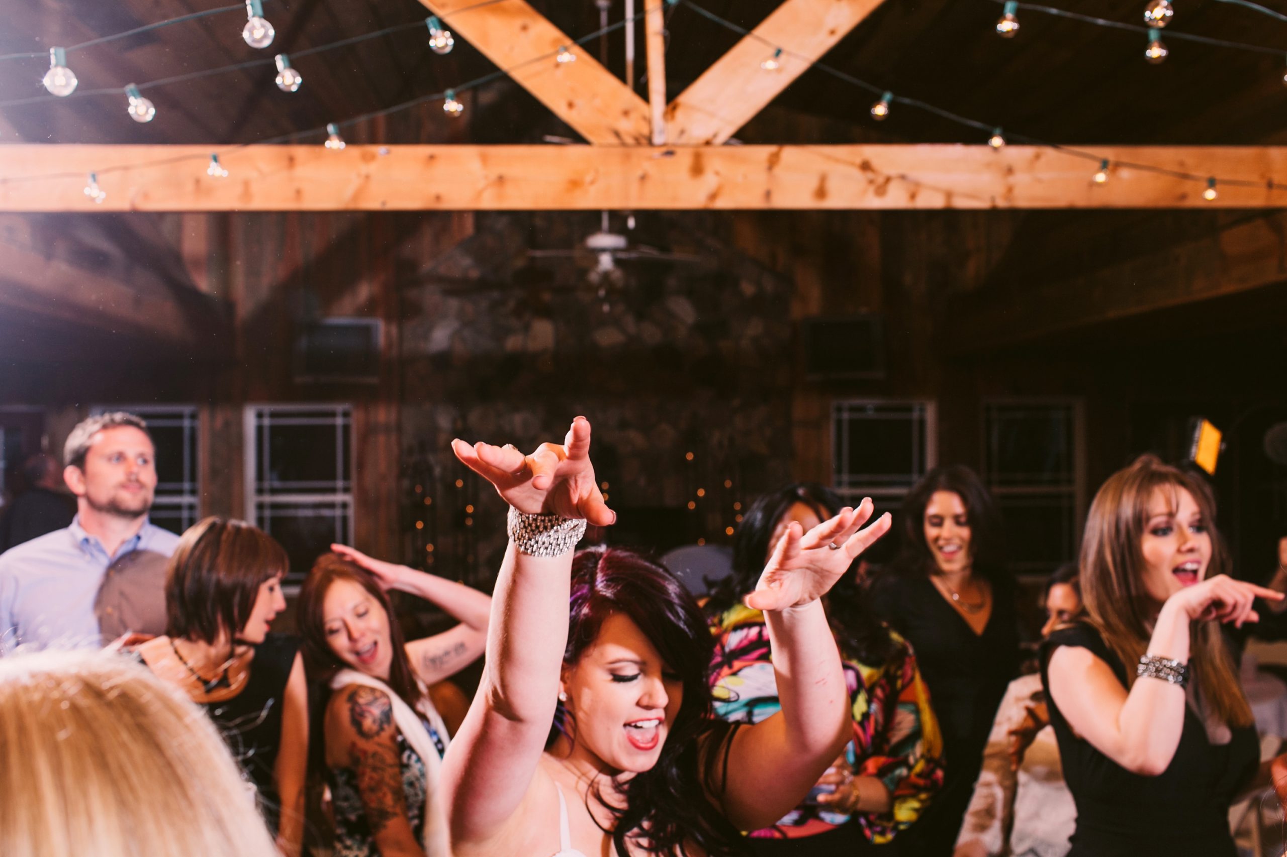 Wedding guests dancing enthusiastically in rustic barn venue with exposed beams and string lights