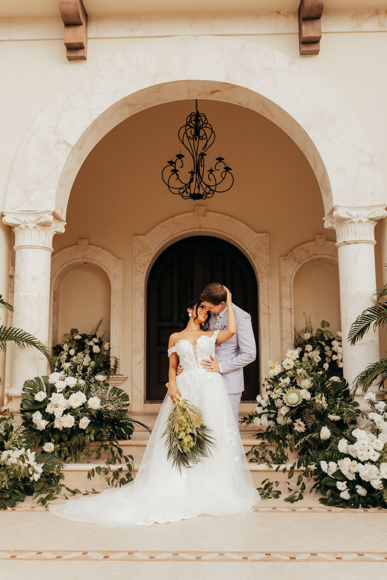 Newlywed couple embracing under ornate archway adorned with white and green floral arrangements