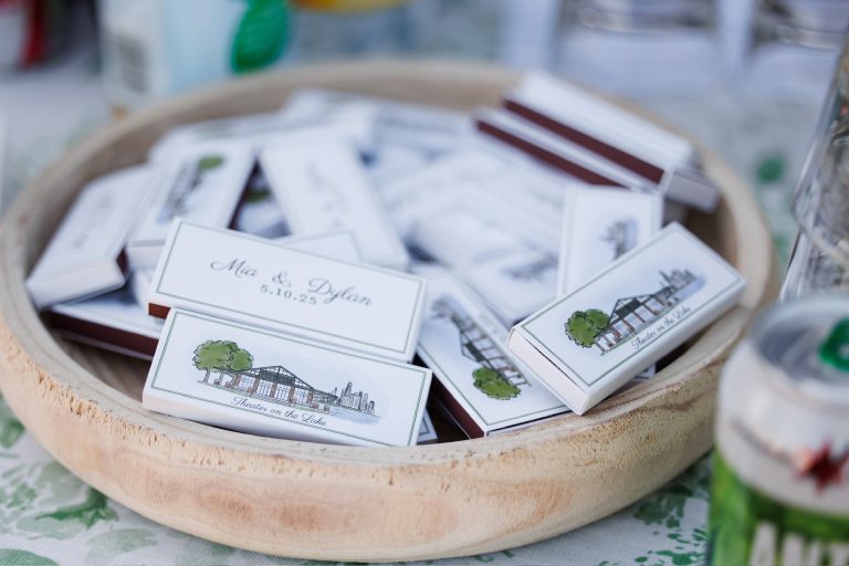 Personalized wedding favor matchboxes in wooden bowl displaying venue illustration and couple's names with date