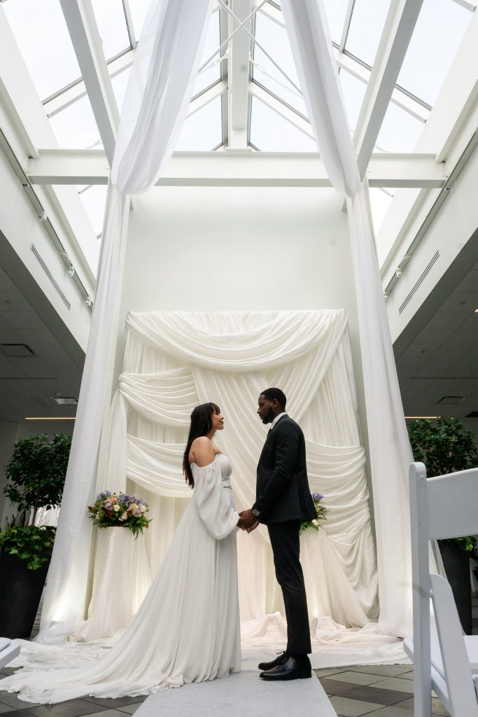 Bride and groom holding hands before draped white fabric ceremony backdrop under skylight with floral arrangements