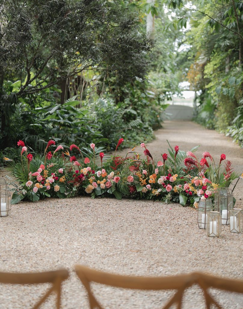 Vibrant tropical floral arrangement with pink and coral blooms lines garden ceremony aisle flanked by lanterns