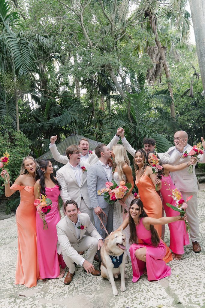 Wedding party in coral and pink attire celebrates with dog in tropical garden setting
