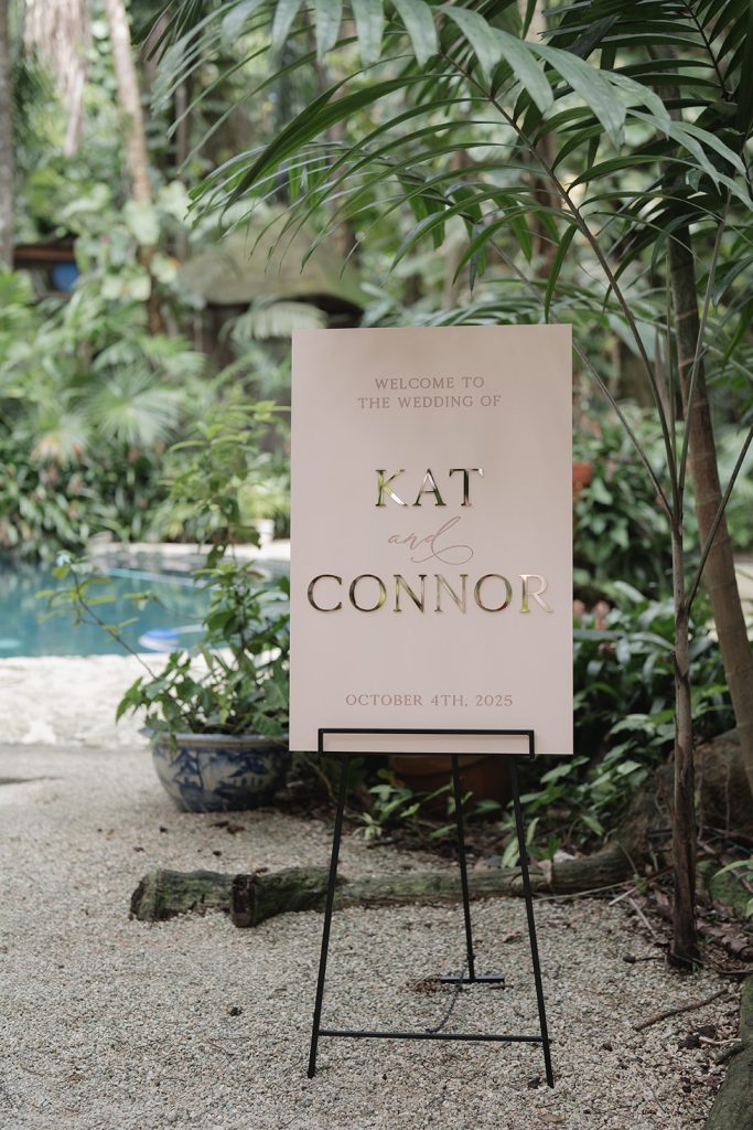Blush wedding welcome sign for Kat and Connor displayed on easel among tropical palm fronds