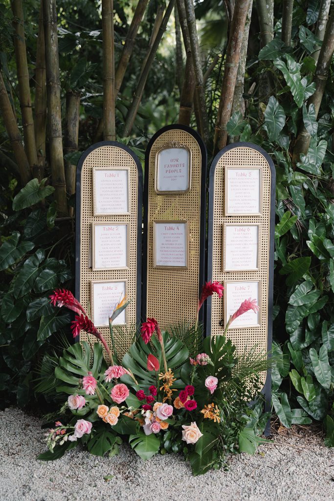 Gold cane-backed seating chart displaying table assignments surrounded by tropical greenery and colorful blooms