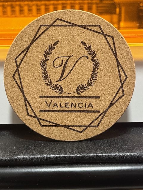 Cork coaster with Valencia monogram featuring letter V in laurel wreath and geometric border