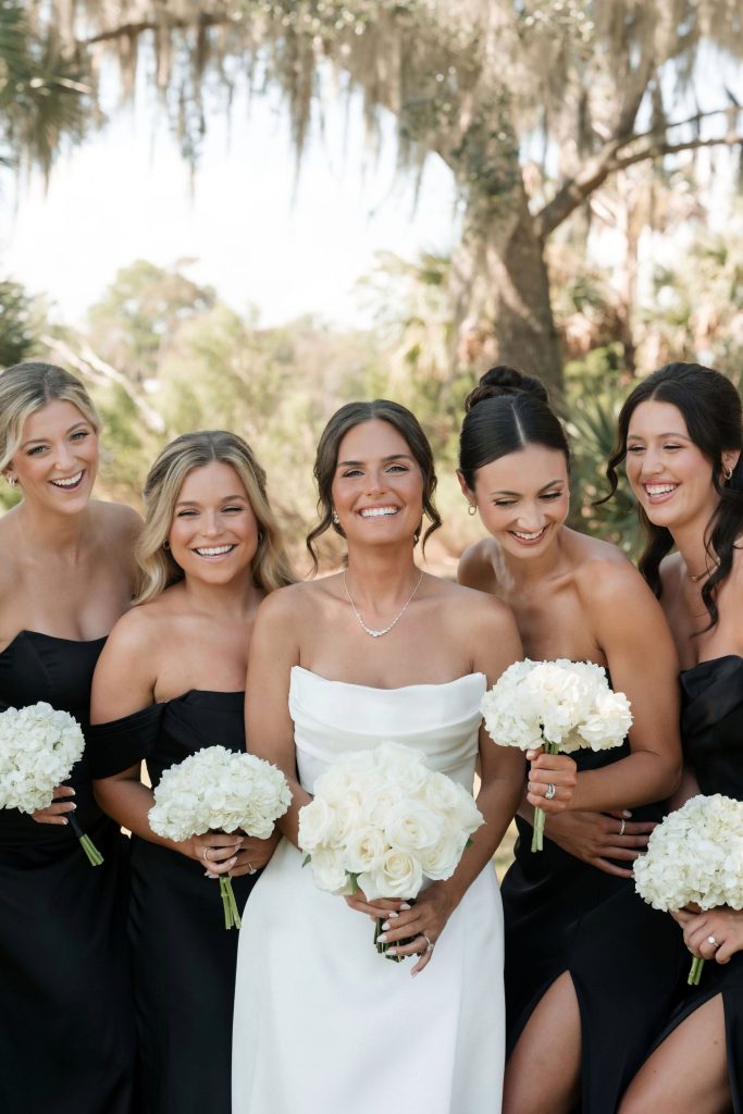 Bride in white strapless gown with bridesmaids in black off-shoulder dresses holding white rose bouquets outdoors under Spanish moss trees