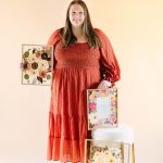 Woman in coral tiered dress holds framed pressed flower keepsakes showcasing various floral preservation styles for wedding mementos