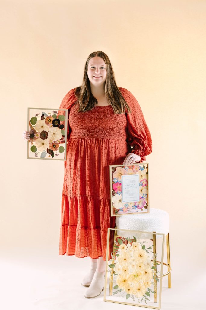 Woman in coral tiered dress holds framed pressed flower keepsakes showcasing various floral preservation styles for wedding mementos