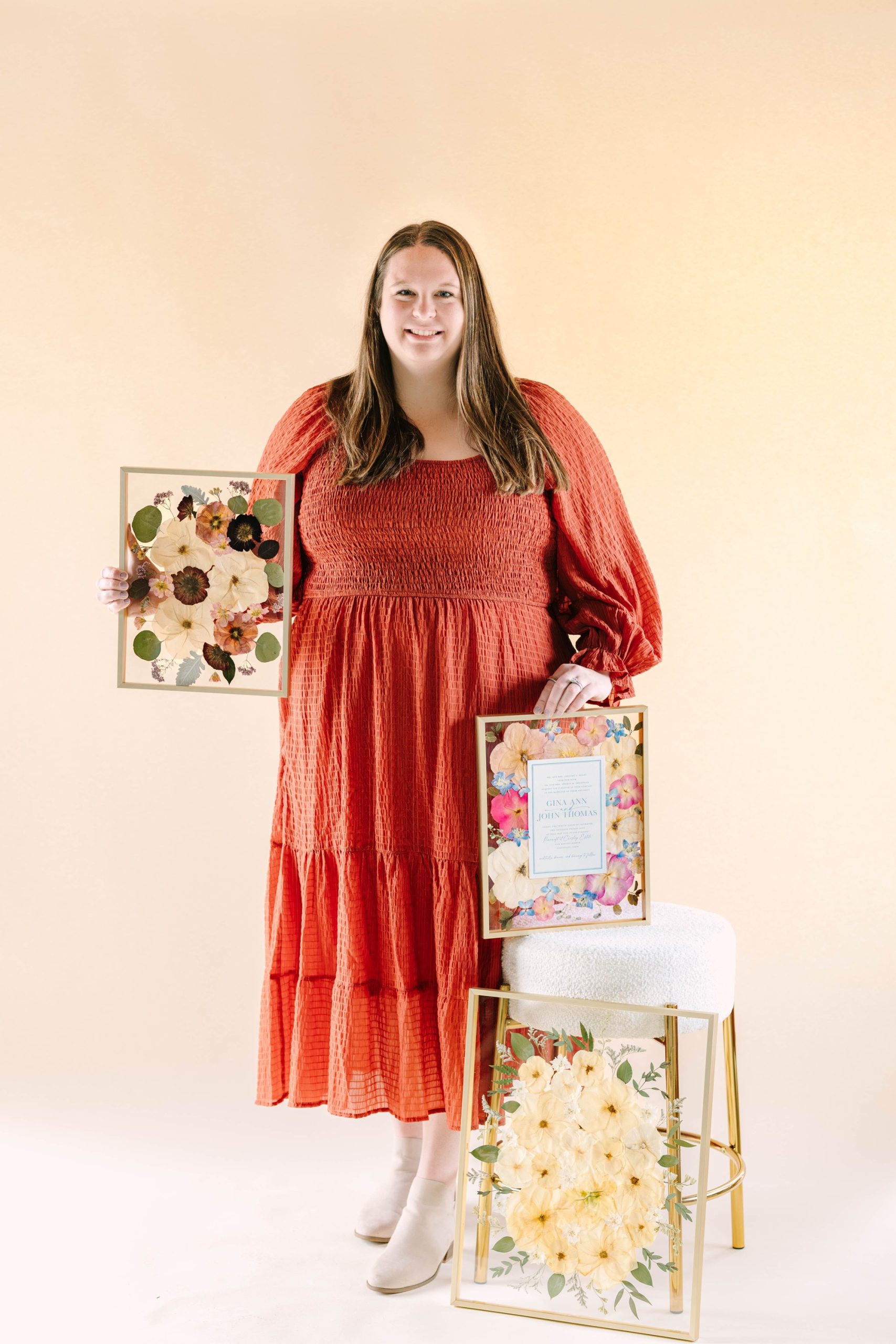 Woman in coral tiered dress holds framed pressed flower keepsakes showcasing various floral preservation styles for wedding mementos