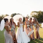 Bride in white gown with bridesmaids in neutral dresses holding baby's breath bouquets and paper parasols on sunny golf course