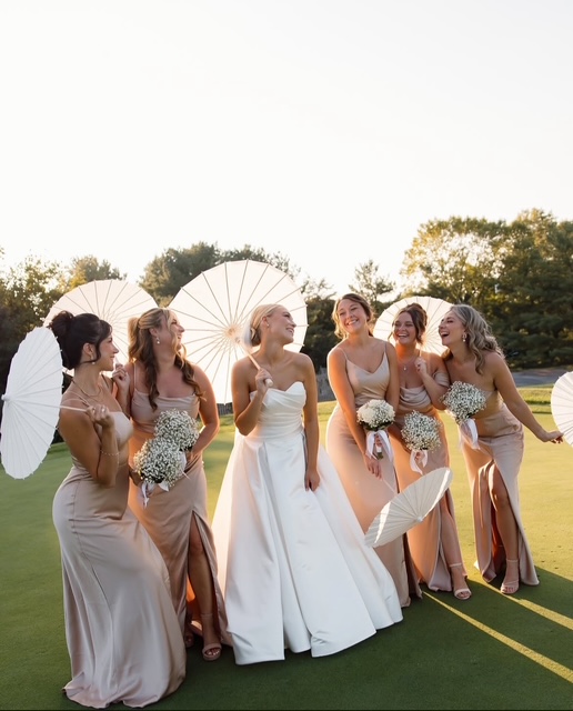 Bride in white gown with bridesmaids in neutral dresses holding baby's breath bouquets and paper parasols on sunny golf course
