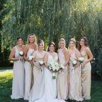 Bride and bridesmaids in neutral-toned gowns holding white bouquets under willow tree