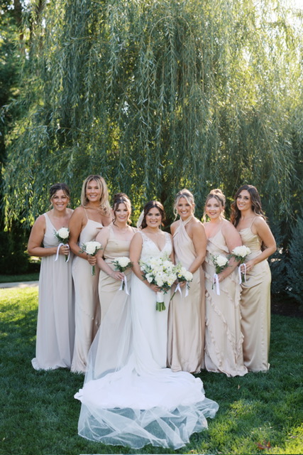 Bride and bridesmaids in neutral-toned gowns holding white bouquets under willow tree