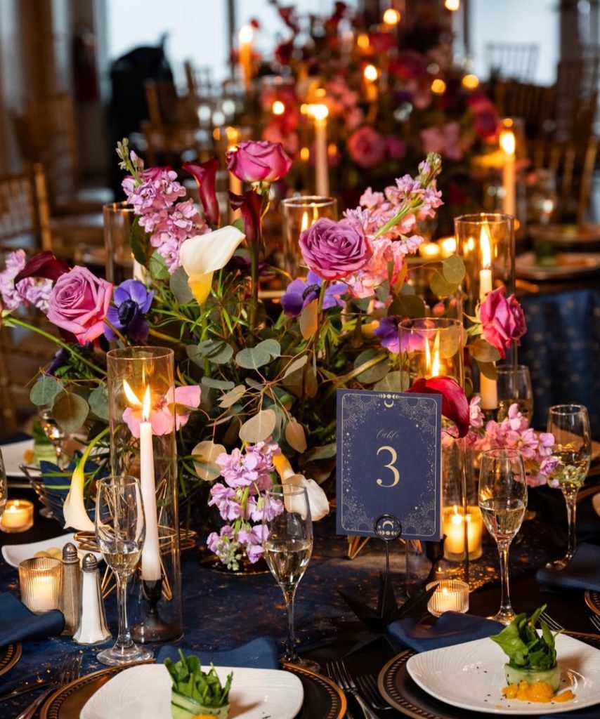 Elegant wedding reception table with purple and pink rose centerpiece, gold candlelight, navy table number three, and formal place settings