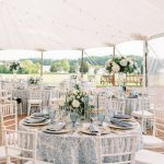 Elegant outdoor wedding reception under white tent with blue floral tablecloths, white chiavari chairs, and lakeside view