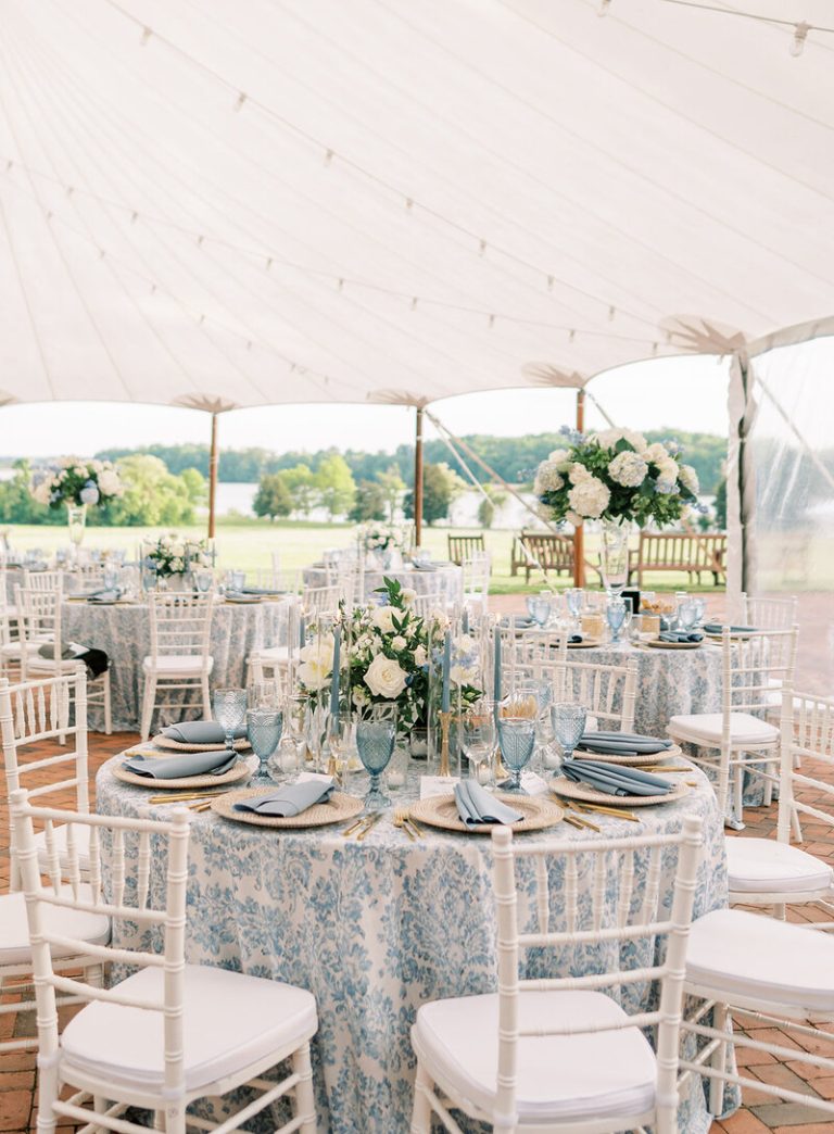 Elegant outdoor wedding reception under white tent with blue floral tablecloths, white chiavari chairs, and lakeside view