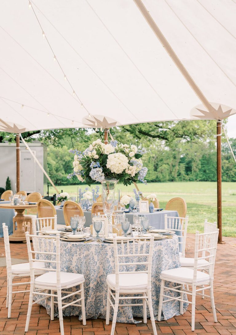 Elegant outdoor wedding reception table under white tent with blue floral linens, tall white hydrangea centerpiece, and white chiavari chairs
