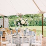 Elegant outdoor wedding reception under white tent with blue floral tablecloth, white chiavari chairs, and tall hydrangea centerpiece