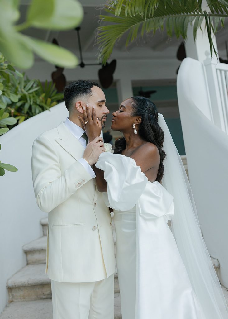 Bride in white gown with dramatic sleeves and groom in cream suit share an intimate moment under tropical palms