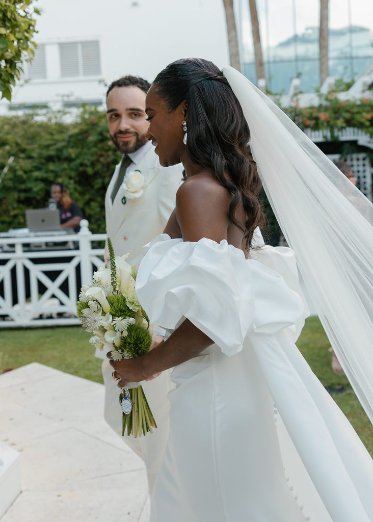 Bride in off-shoulder gown with dramatic sleeves and cathedral veil holds white bouquet beside groom in ivory jacket