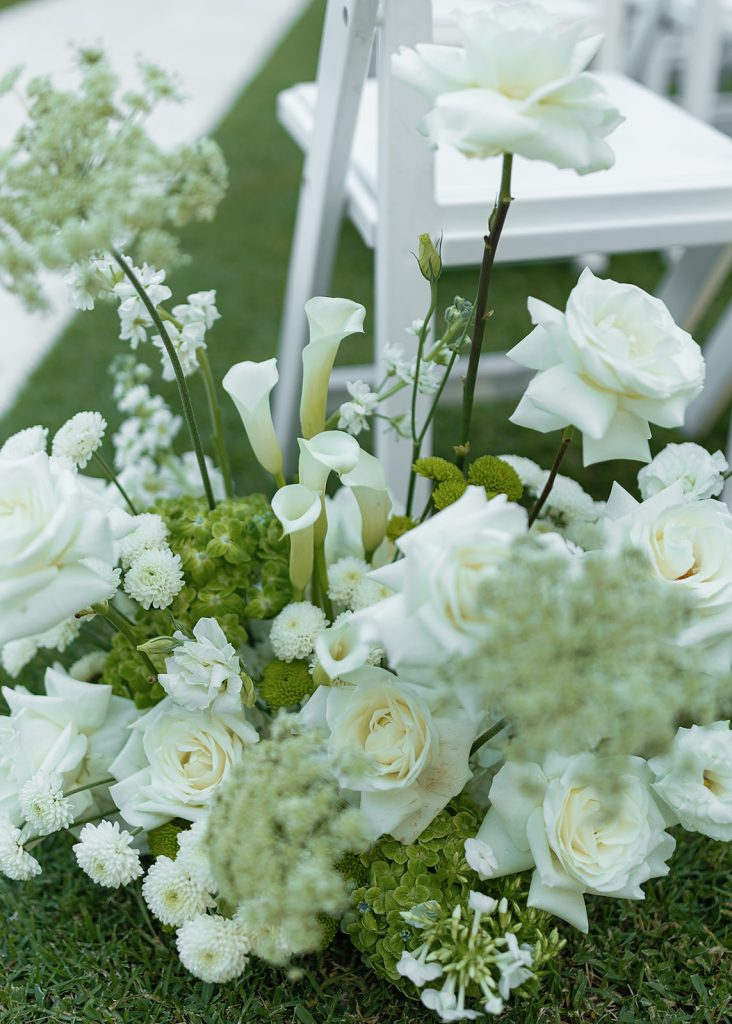 Elegant white and green floral arrangement with blooms and calla lilies beside white ceremony chairs