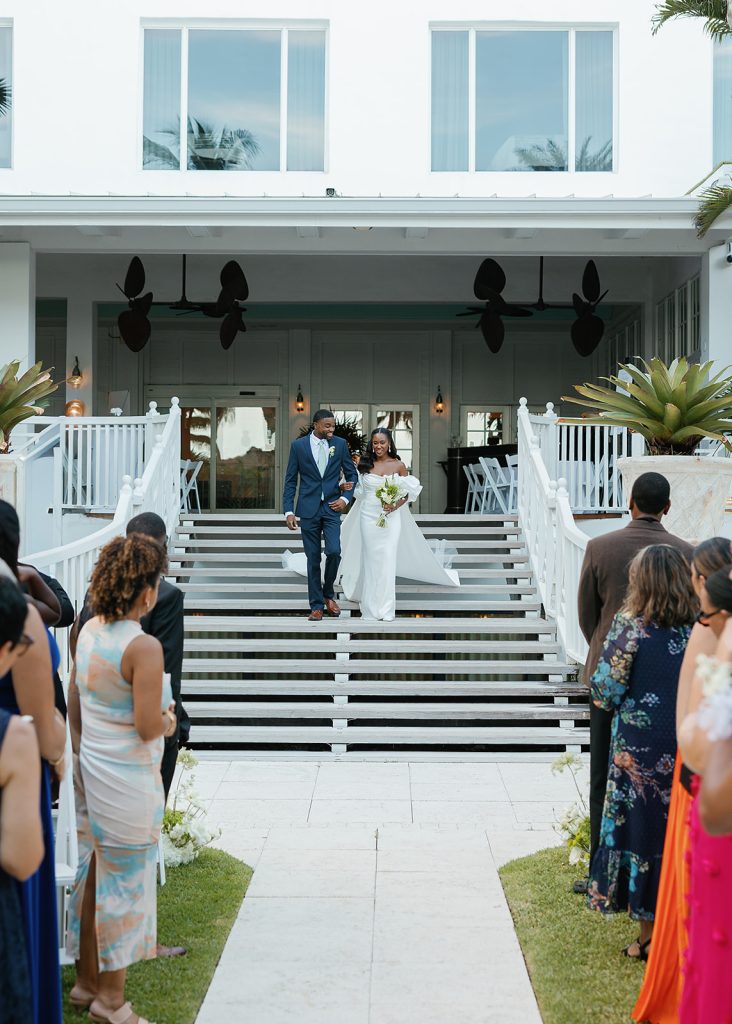 Bride and groom descending white stairs at waterfront venue as guests watch outdoor ceremony
