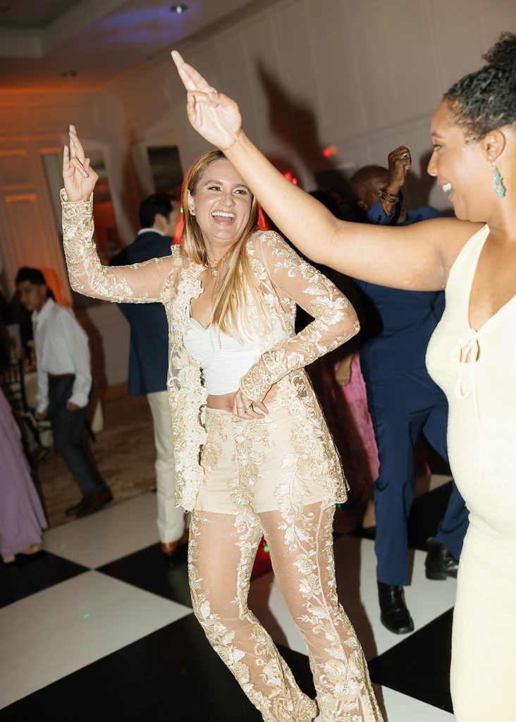 Bride in embroidered lace pantsuit dancing joyfully with guest on black and white checkered dance floor