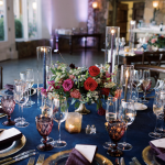 Navy blue tablescape with red and pink rose centerpiece, gold chargers, purple napkins, amber goblets, and tall candle holders