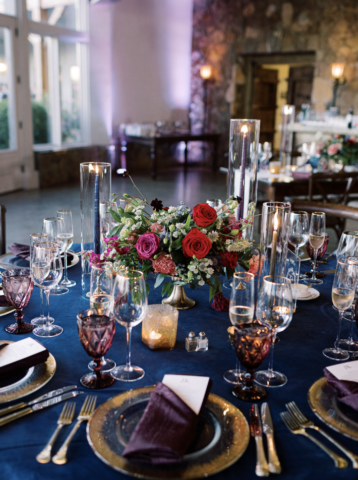 Navy blue tablescape with red and pink rose centerpiece, gold chargers, purple napkins, amber goblets, and tall candle holders