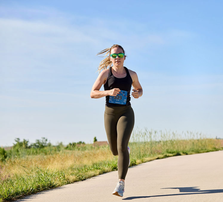 Fitness professional Madeline running in race bib on outdoor trail under clear blue sky