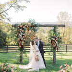 Bride and groom kissing under rustic wooden arbor adorned with pink and coral florals at outdoor Northern Virginia wedding