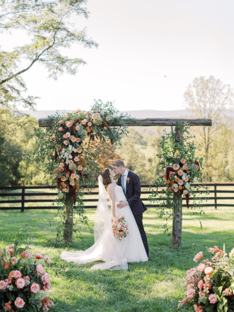 Bride and groom kissing under rustic wooden arbor adorned with pink and coral florals at outdoor Northern Virginia wedding