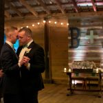 Two grooms share first dance in rustic barn venue with string lights