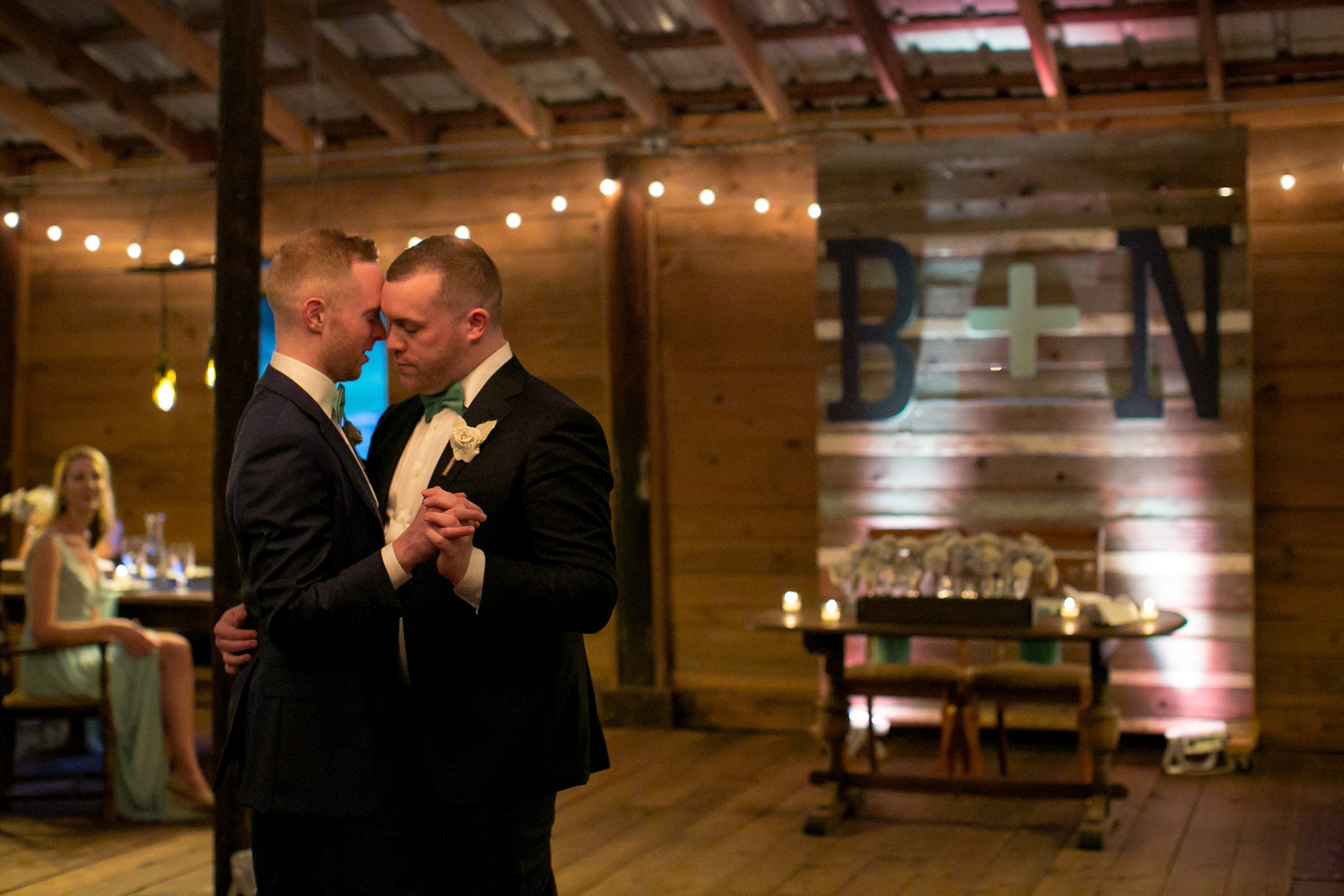 Two grooms share first dance in rustic barn venue with string lights