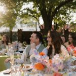 Wedding guests seated at outdoor reception table decorated with floral centerpieces at sunset