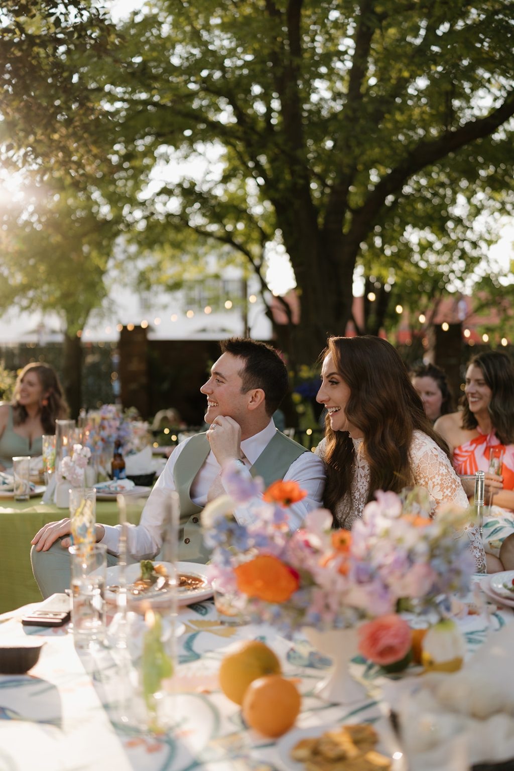 Wedding guests seated at outdoor reception table decorated with floral centerpieces at sunset
