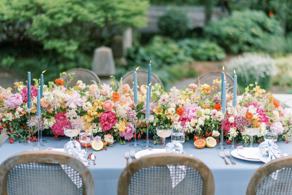 Outdoor garden wedding reception table with vibrant pink, yellow, and orange floral centerpieces and blue taper candles