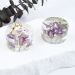 Two clear resin keepsakes containing preserved purple wedding flowers and gold jewelry on white surface