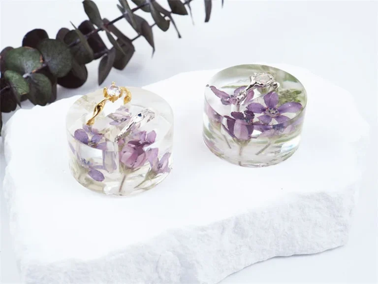 Two clear resin keepsakes containing preserved purple wedding flowers and gold jewelry on white surface