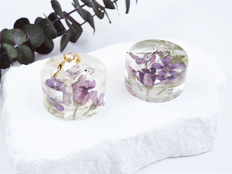 Two clear resin keepsakes containing preserved purple wedding flowers and gold jewelry on white surface