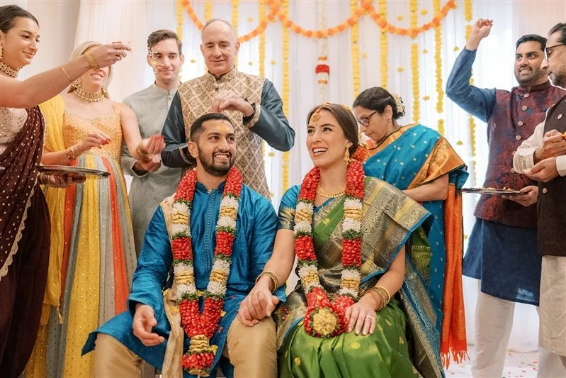 Bride and groom in traditional Indian wedding attire seated during ceremony with family showering them with flower petals