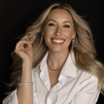 Woman in white shirt with long blonde hair and jewelry smiling at camera on black background