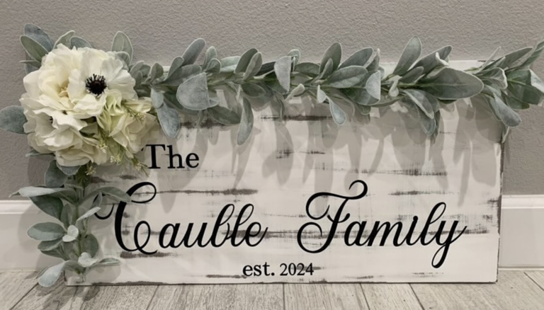 Rustic wooden sign reading "The Cauble Family est. 2024" adorned with eucalyptus garland and white roses