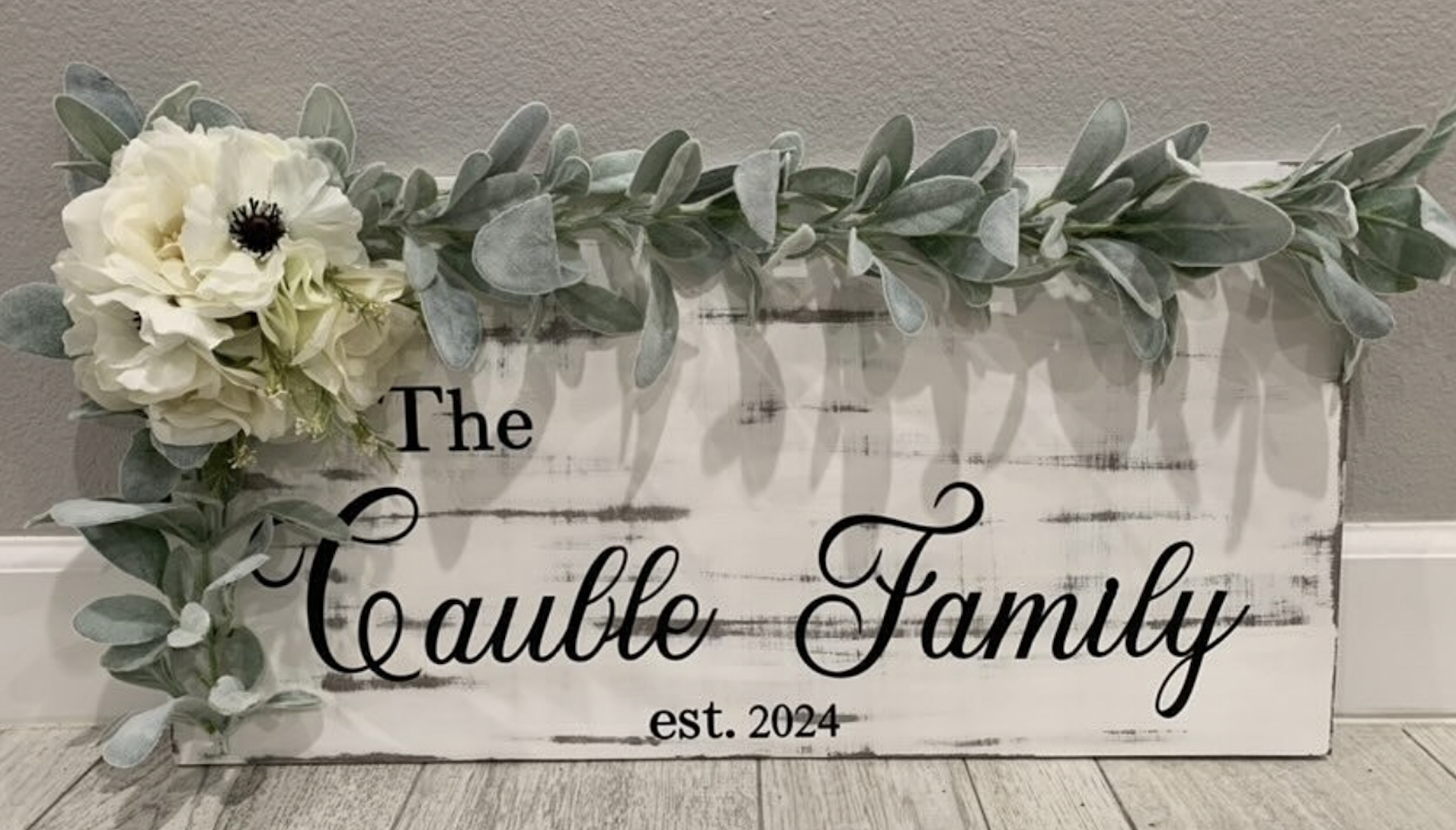 Rustic wooden sign reading "The Cauble Family est. 2024" adorned with eucalyptus garland and white roses