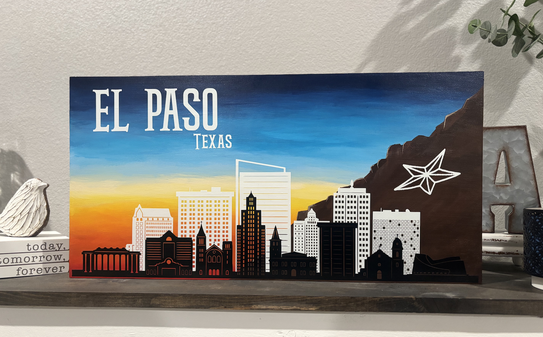 El Paso Texas skyline painting featuring downtown buildings, Franklin Mountains, and star logo against blue and orange sunset sky