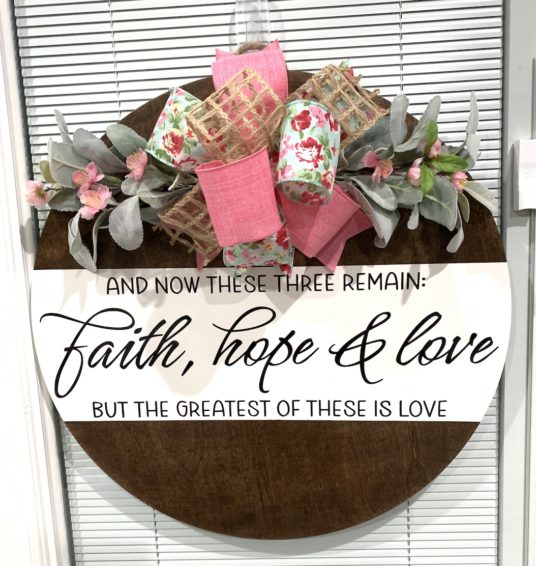 Round wooden door hanger with pink and floral ribbons, eucalyptus leaves, and "Faith, hope & love" biblical quote