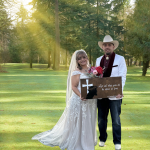 Bride in white lace gown with veil and groom in cowboy hat holding scripture sign on sunlit golf course