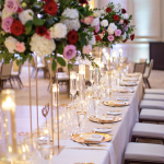 Elegant wedding reception table with tall rose centerpieces, gold-rimmed place settings, and pillar candles