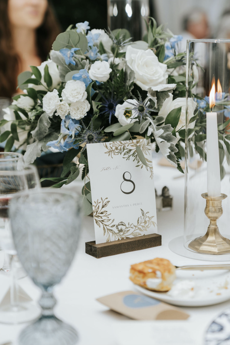 Wedding table centerpiece with white roses, blue flowers, table number 8 card, and lit candles