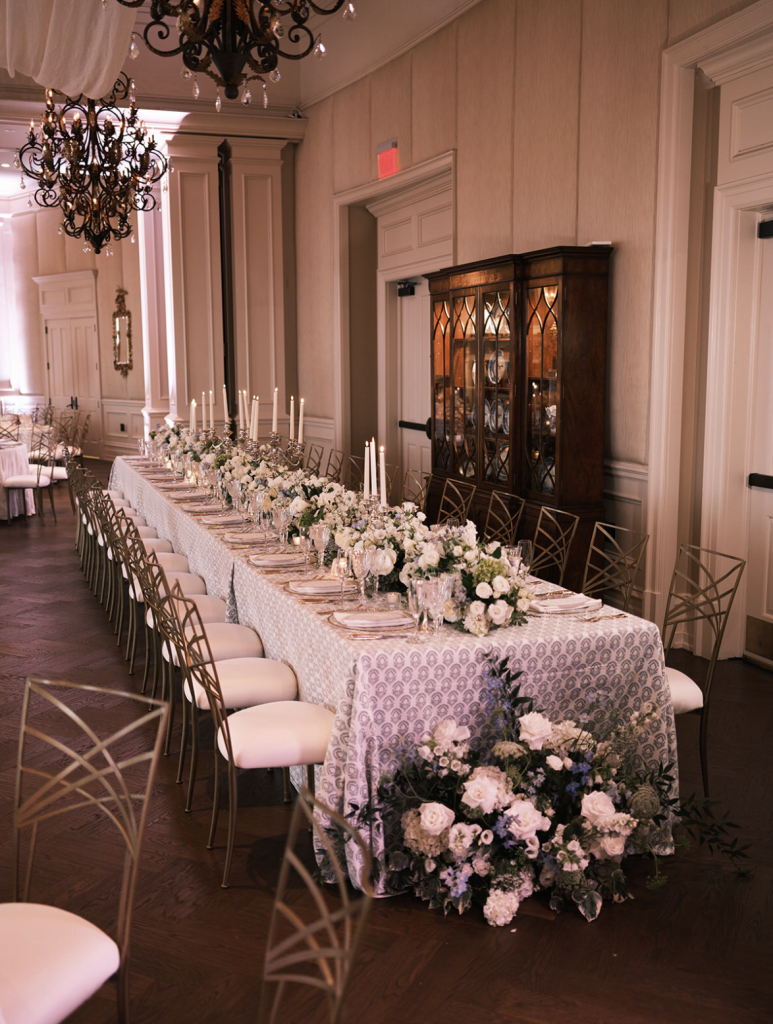 Elegant long reception table with white floral centerpieces and gold chairs in ballroom with chandelier and antique cabinet